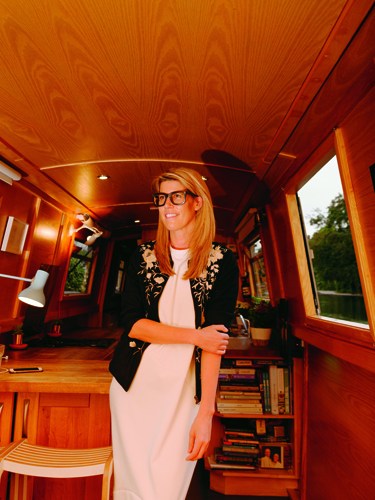 Woman standing inside a wooden-lined mobile space with shelves of books and warm orange lighting.