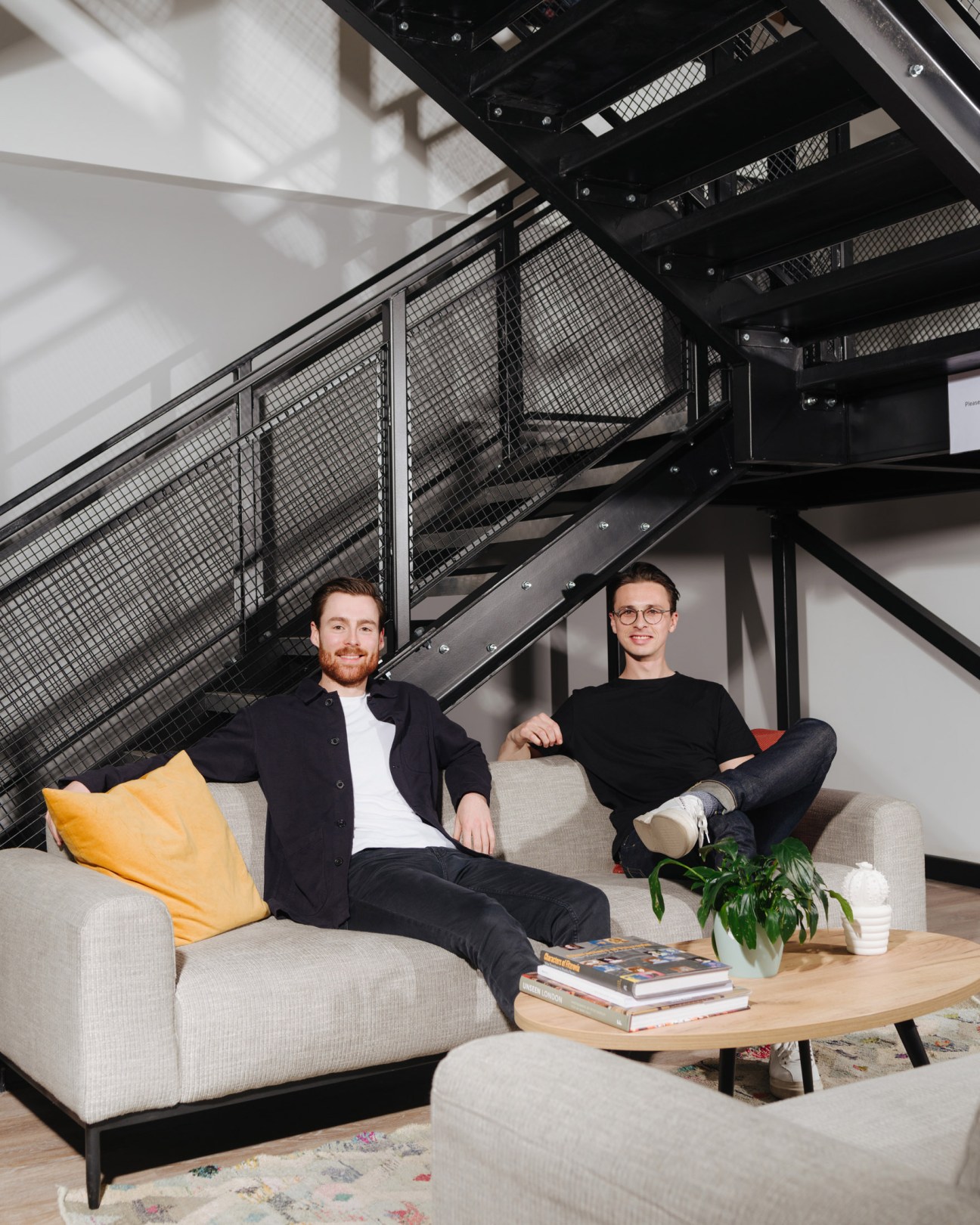 Two men sit on a beige sofa in a modern loft space with industrial black staircase and mesh railing behind them.