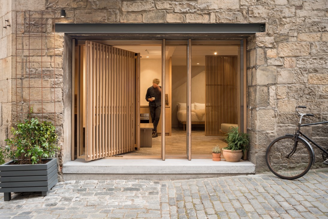 Minimalist studio entrance with wooden slat door open, revealing interior with person inside, stone facade, bicycle parked outside.