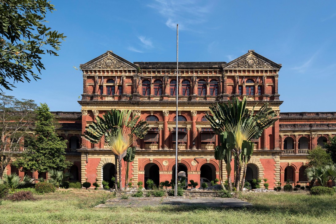 A restored red-brick colonial building with ornate architectural details and palm trees in front under blue sky.
