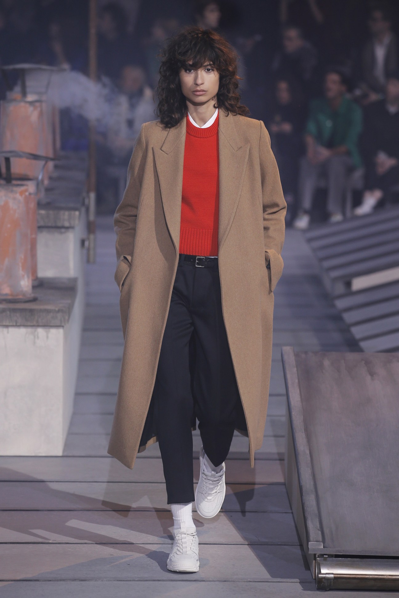Model wearing Ami women's shirt, sweater and double-breasted coat with men's trousers and trainers on runway.