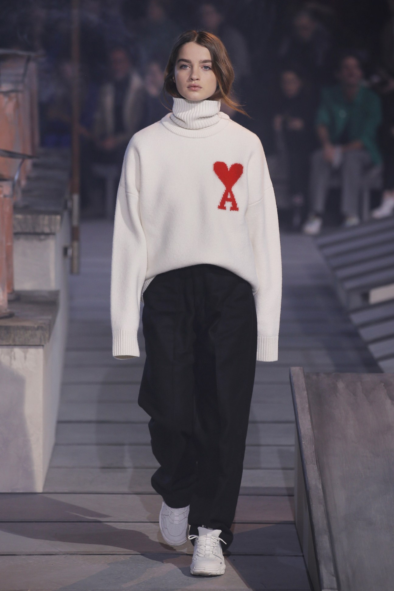 Model wearing Ami white turtleneck sweater with red heart logo and black trousers on runway.