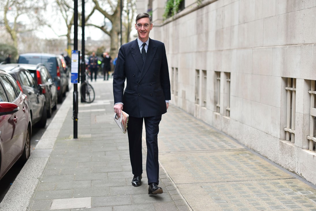 Man in dark double-breasted coat and striped shirt walking on London street carrying publication.