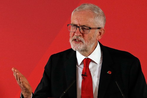 Man in black suit and red tie speaking at a podium against a red background.