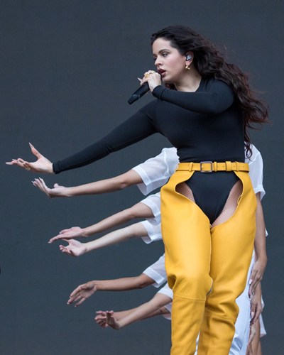 Female performer on stage with microphone wearing black long-sleeved top and yellow pants with gold belt.