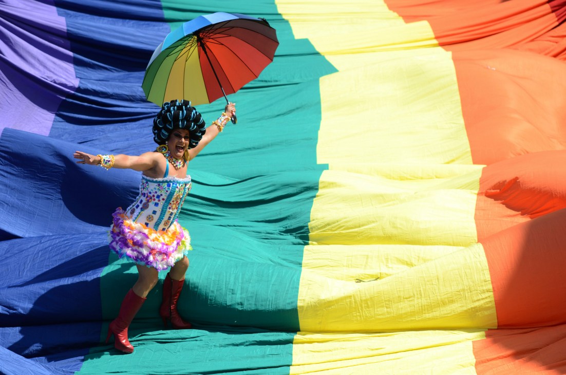 Person in colorful carnival costume with rainbow umbrella against pride flag backdrop.