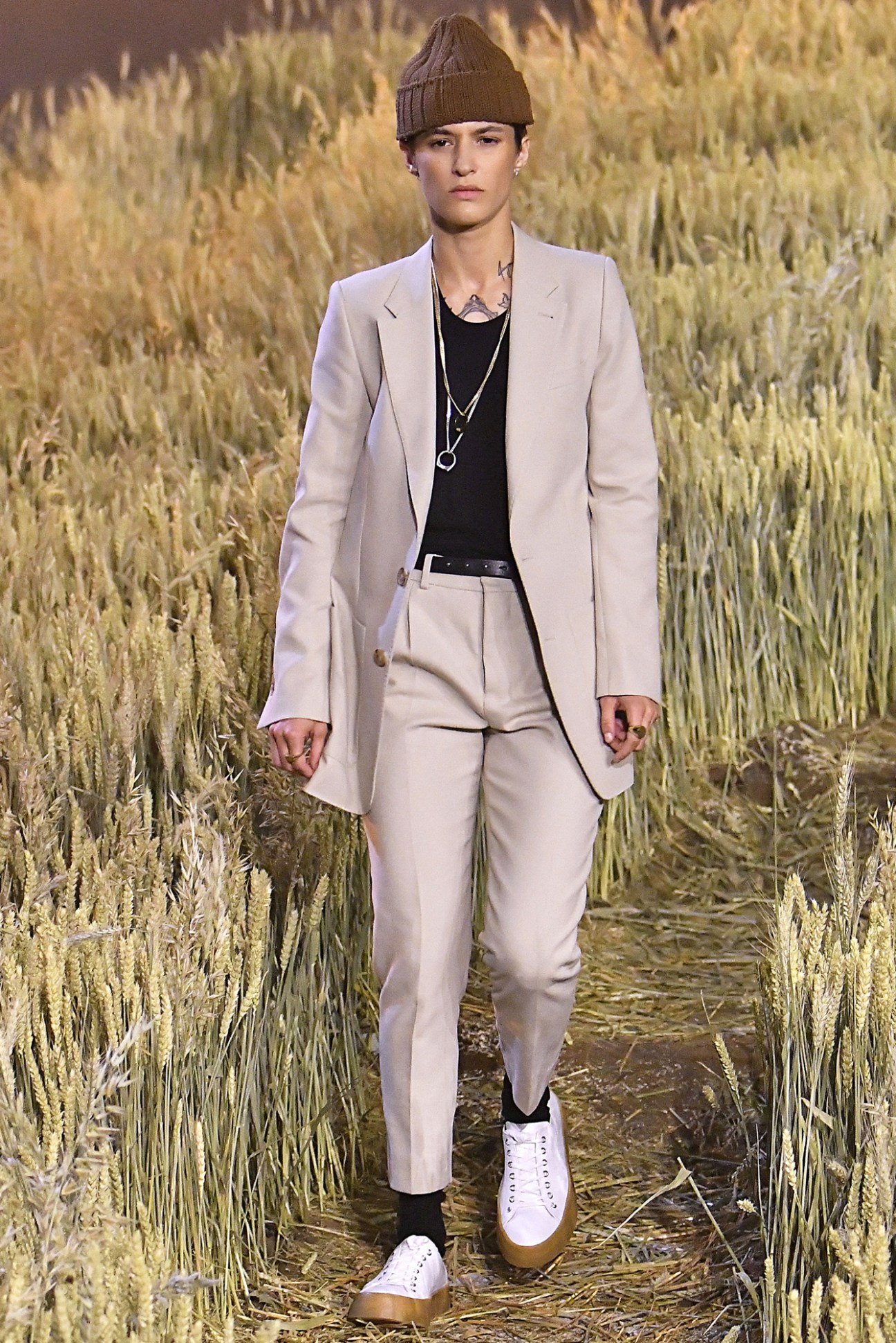 Model wearing Ami women's long jacket and trousers with men's ribbed beanie and trainers on a runway.