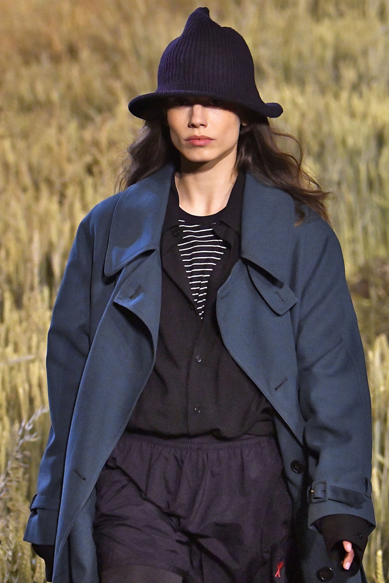 Model wearing women's trench coat, men's striped T-shirt, wide-fit shirt, swim shorts and rib-knitted bucket hat from Ami.