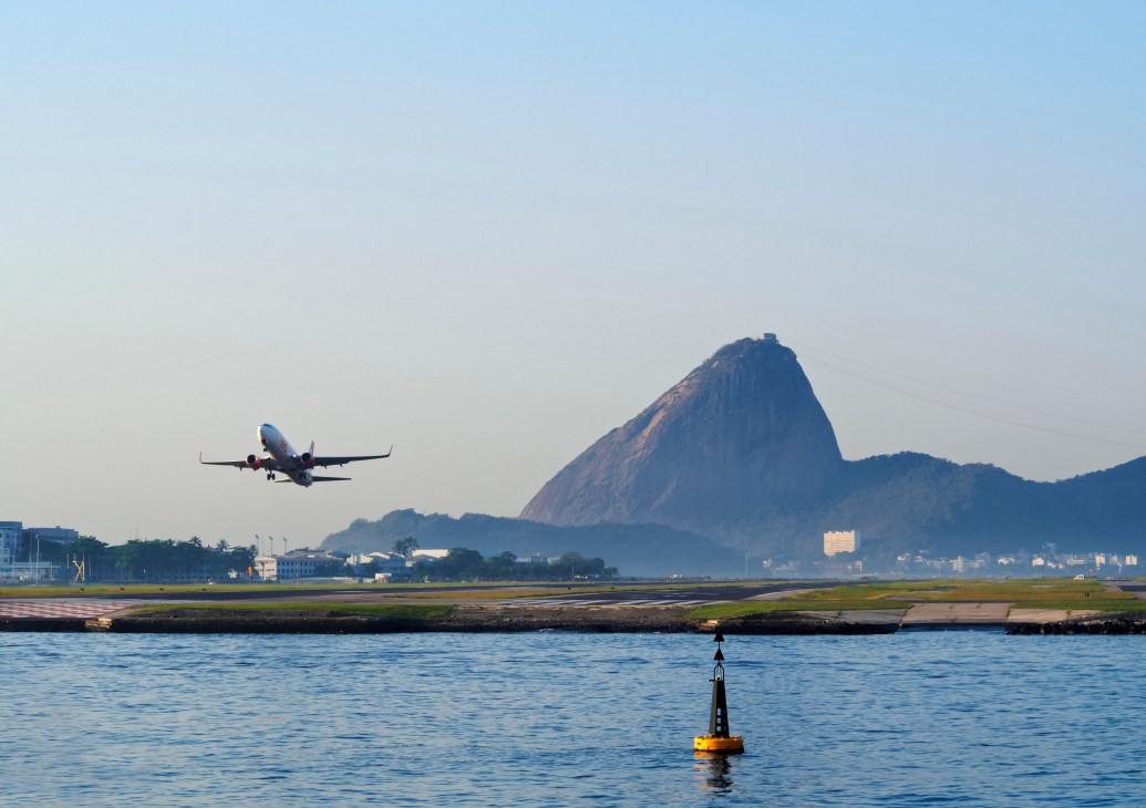 Airplane taking off over water with Sugar Loaf Mountain visible in Rio de Janeiro.