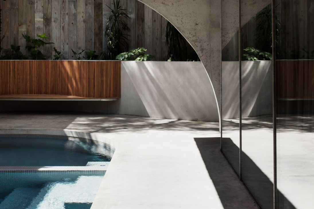 Concrete poolside with timber bench, glass panels, and planted areas in modern residential setting.