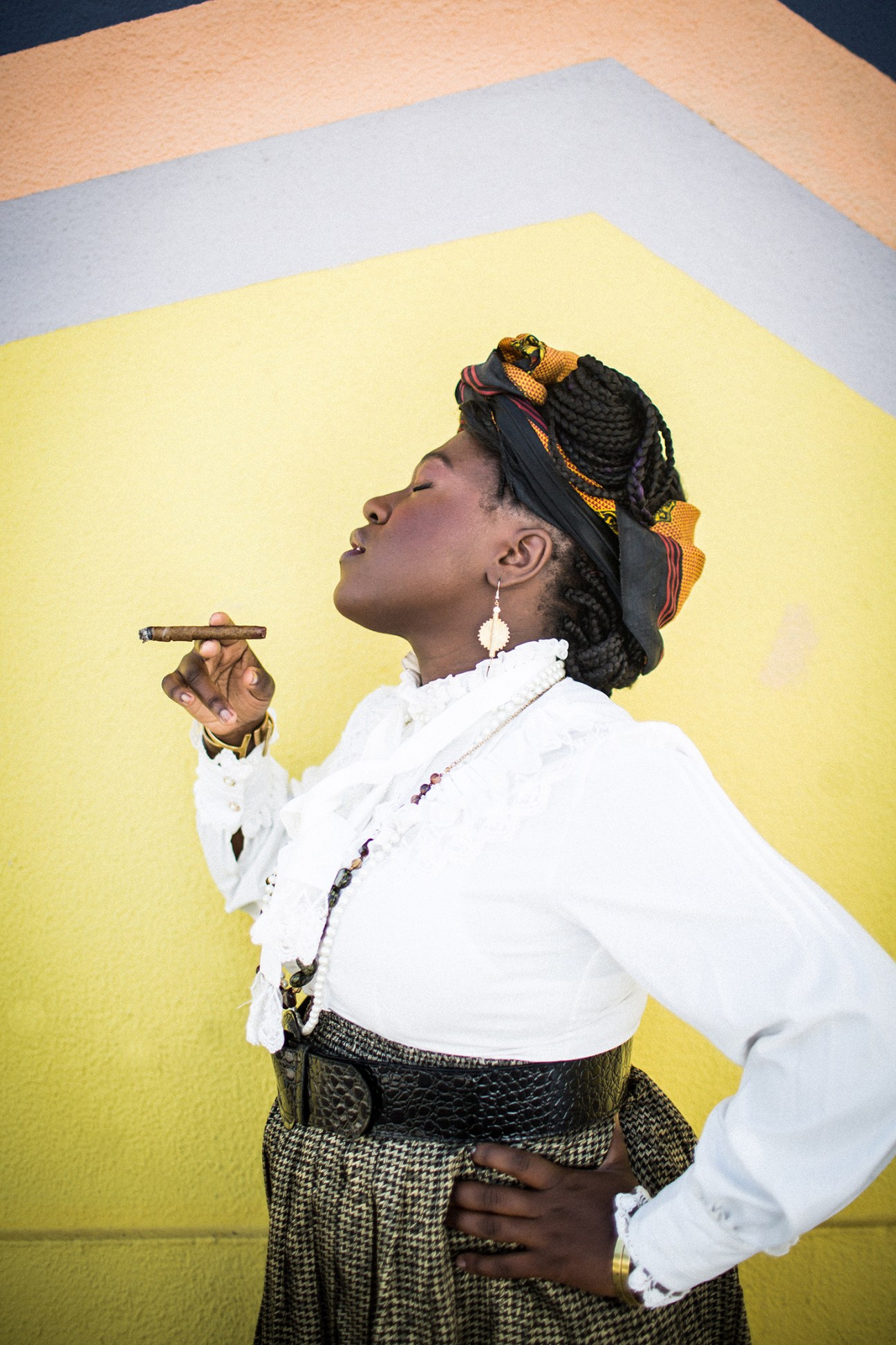Woman in white shirt and patterned skirt holding a cigar, photographed against a yellow and blue background.