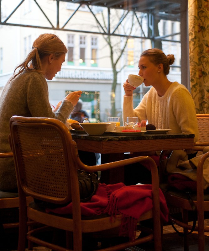 Two women sit at a table by a large window in Café Ekberg, eating pastries and drinking coffee.