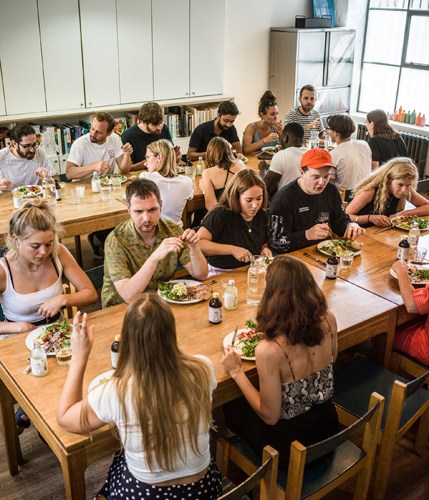 Staff members gathered around wooden tables eating lunch together in a bright office space with shelving in the background.