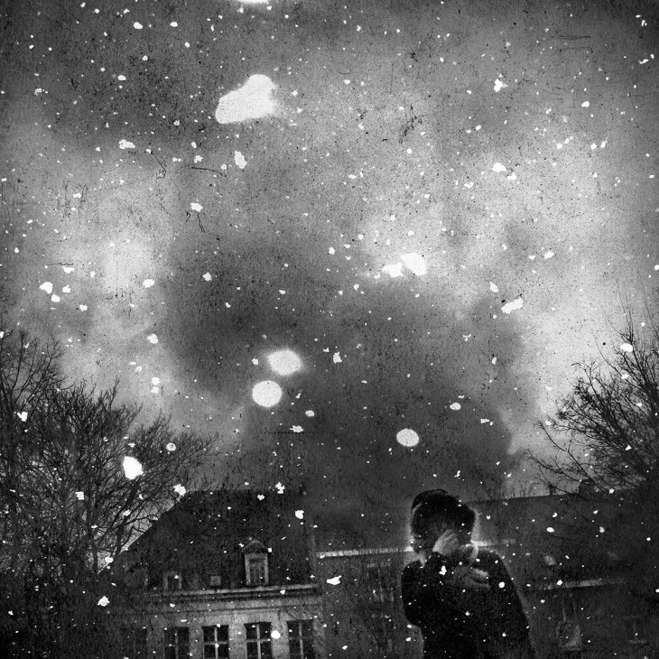 Black and white composite image of a snowy night sky above a building with bare trees, by Hideyuki Ishibashi.