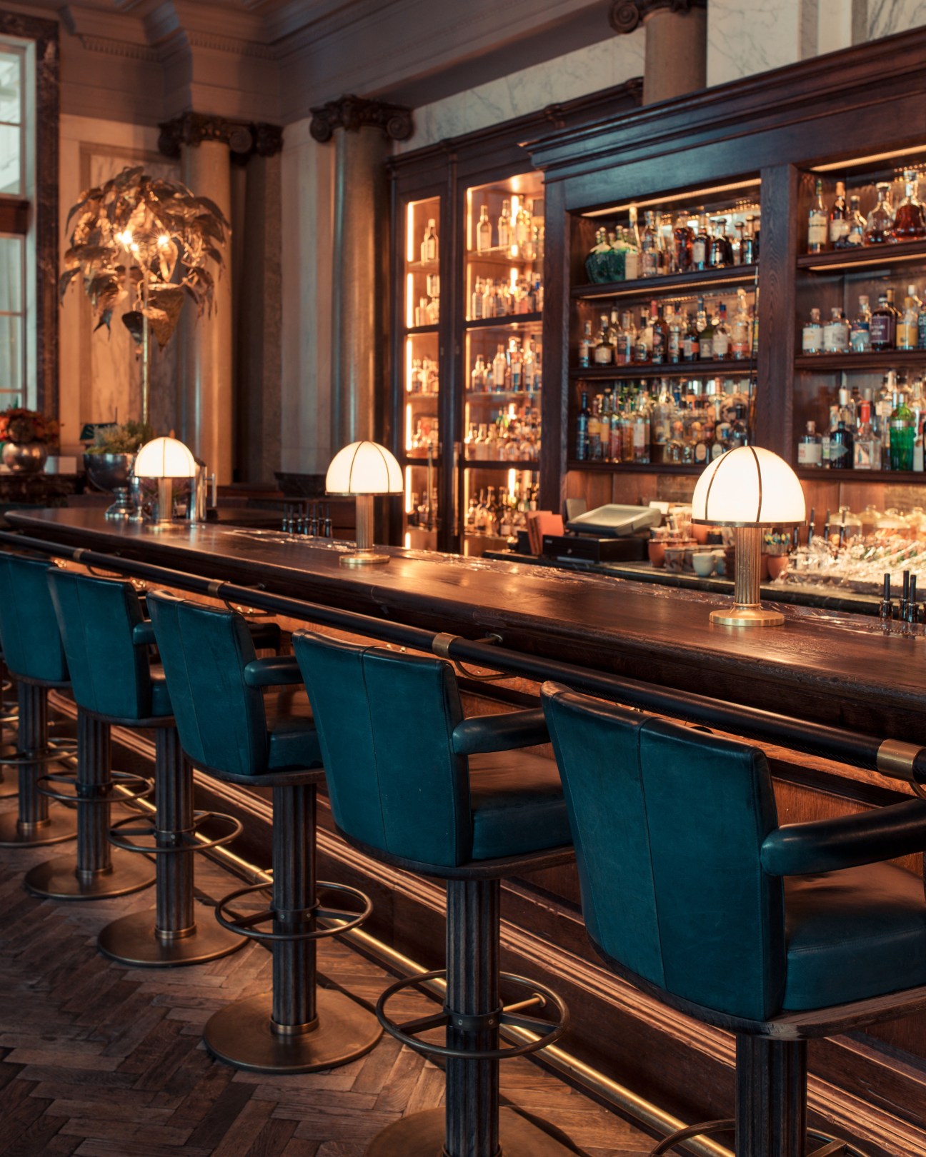 Scarfes Bar at the Rosewood Hotel with illuminated display shelves of spirits and dome-shaped pendant lamps over a dark wooden bar counter.