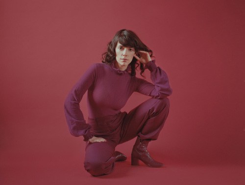 Portrait of Natalie Prass crouching against a red background wearing purple clothing.
