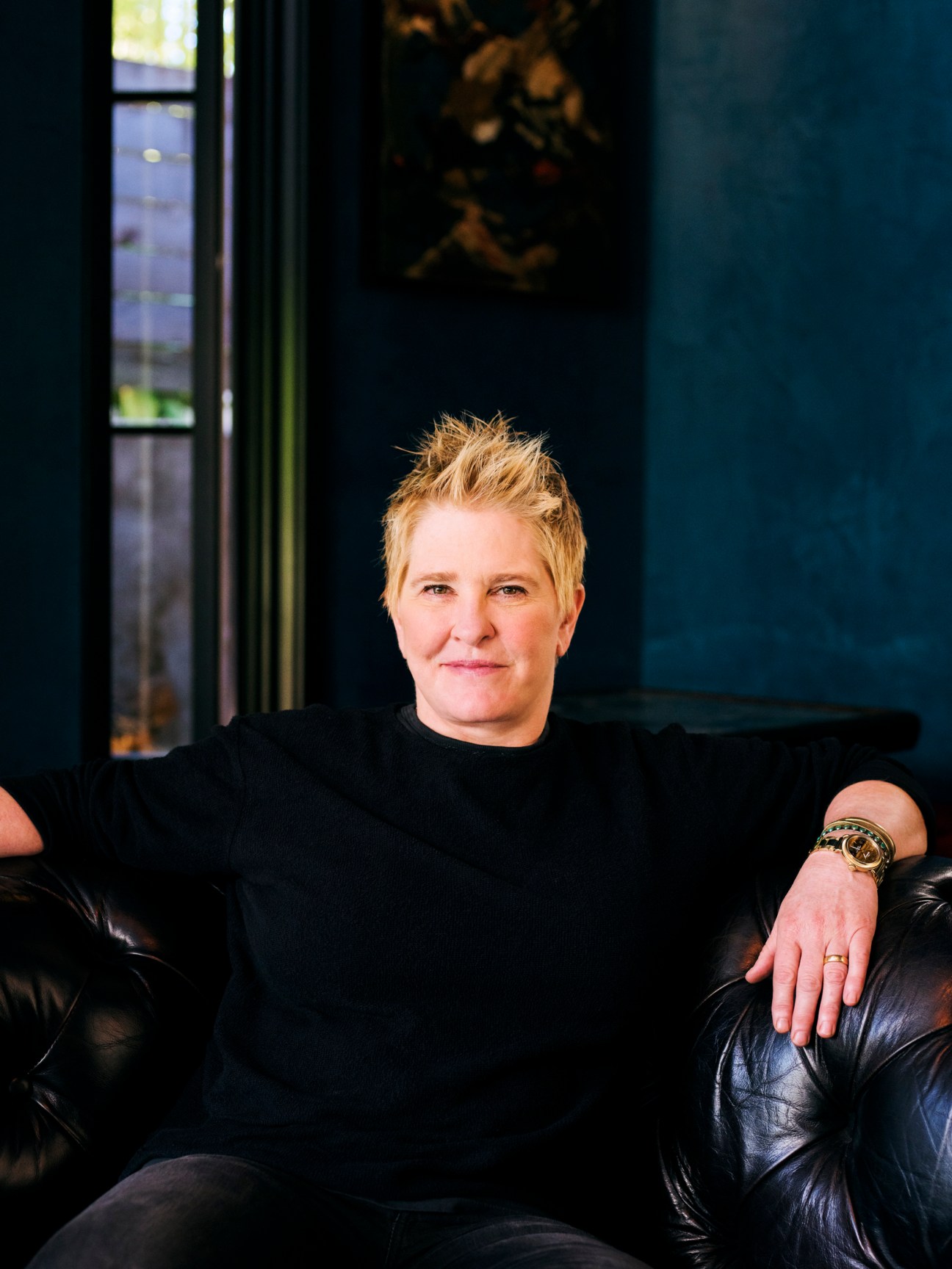 Liz Lambert sitting on black leather sofa in dark interior space wearing black clothing