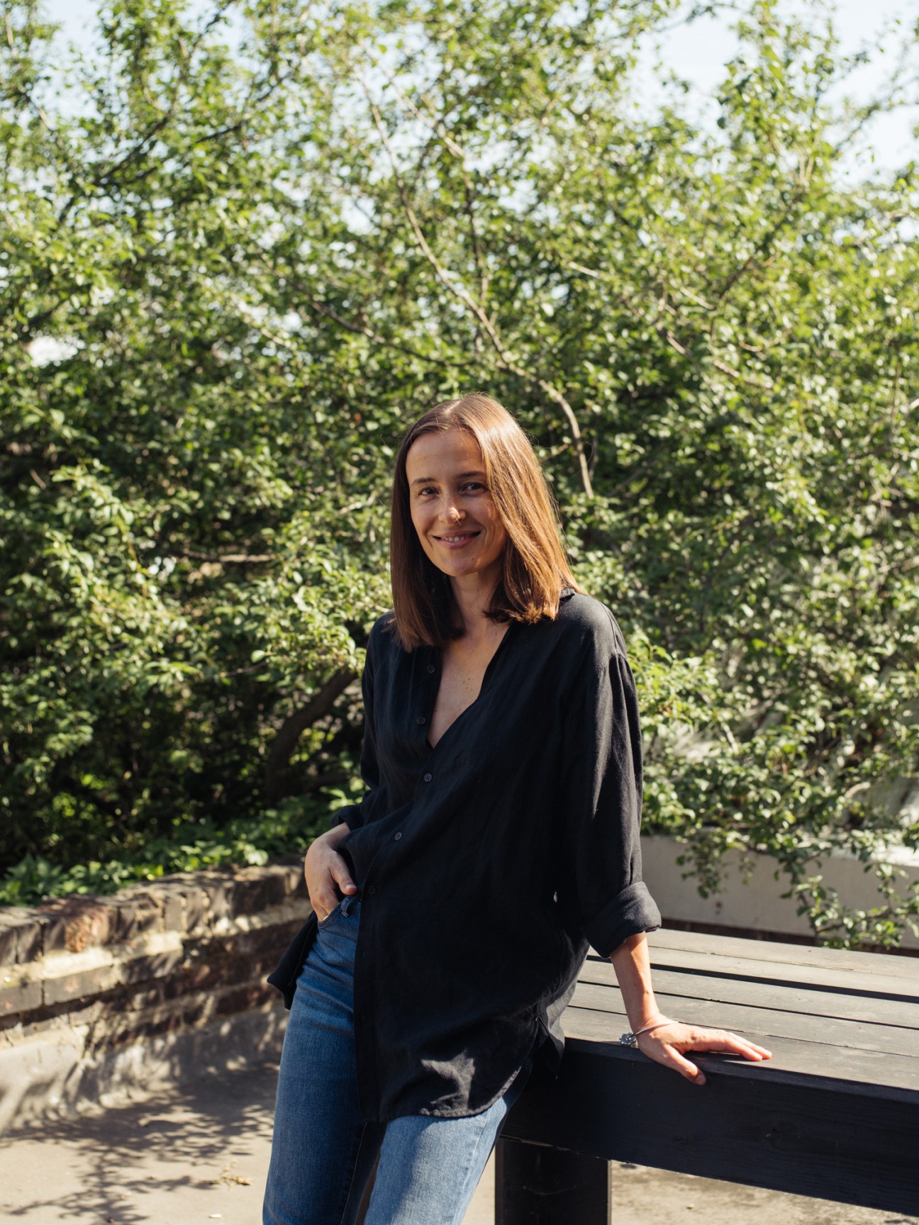 Woman in black shirt and jeans standing in front of lush green foliage, smiling at camera.