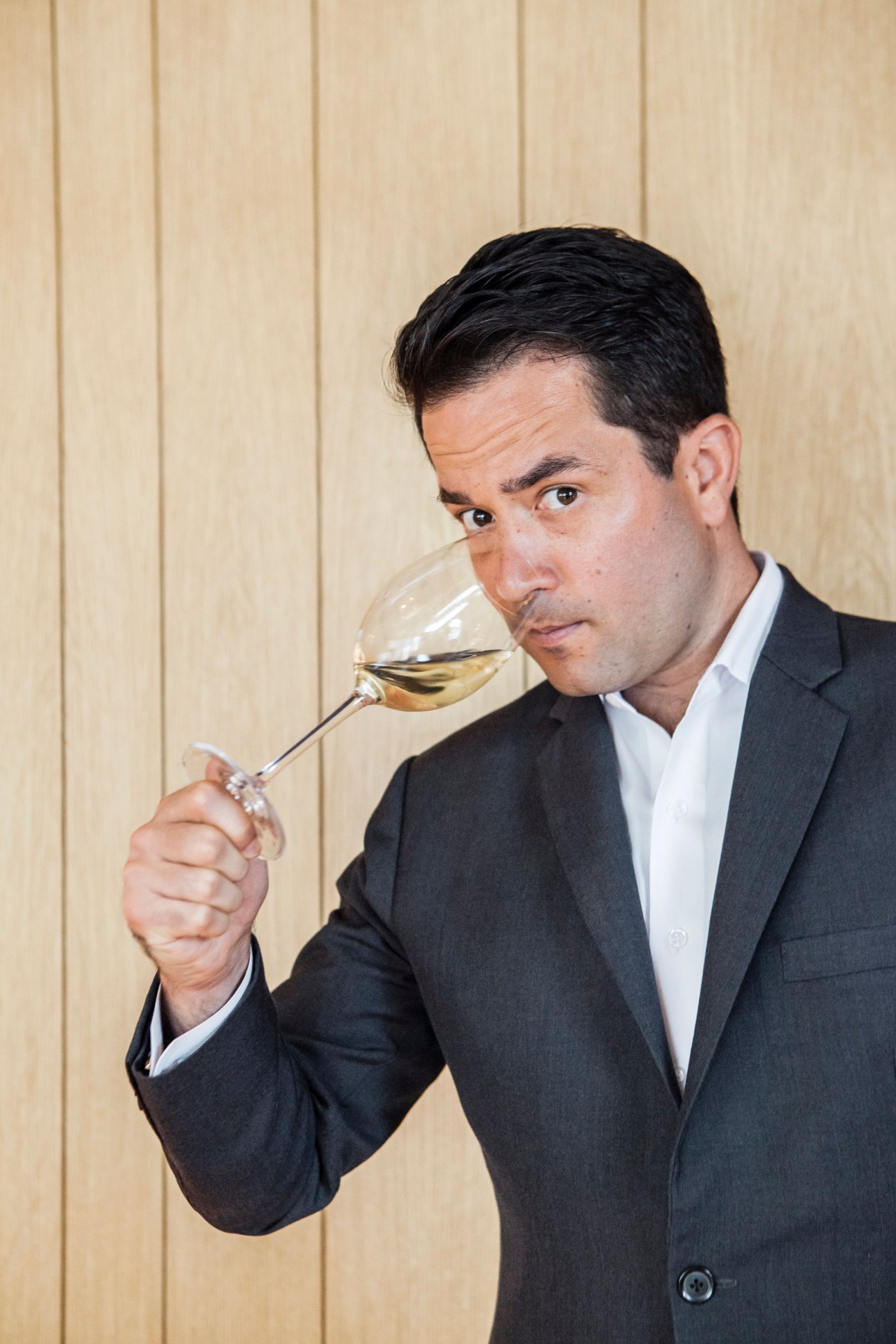 Man in dark suit tasting white wine from a glass against a beige paneled wall.