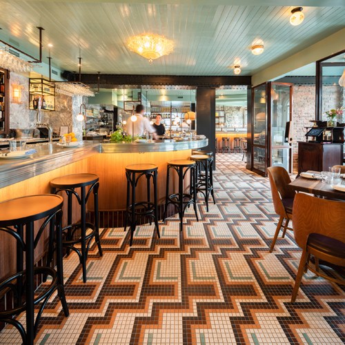 Interior of Un Po' Di Più restaurant in Montreal with orange bar counter, black stools, patterned tile flooring, and open kitchen.