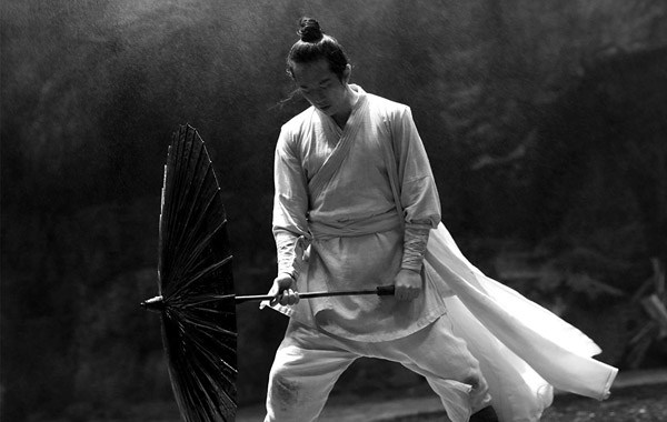 Black and white photograph of a man in traditional white robes wielding a large circular fan-shaped weapon in a martial arts stance.