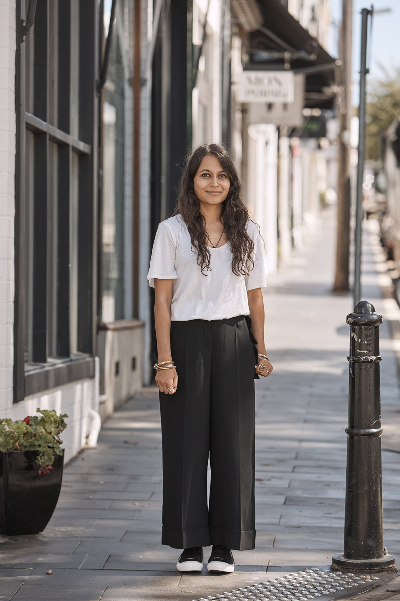 Woman in white t-shirt and black trousers standing on a tree-lined city street.