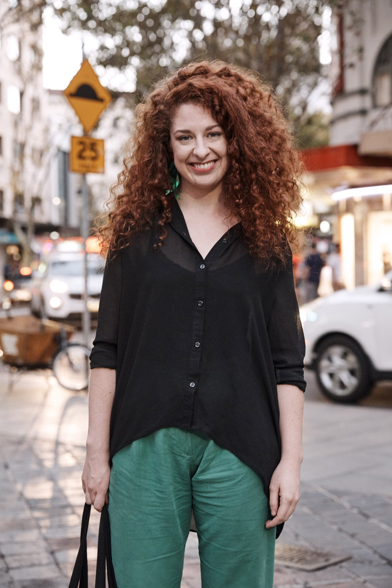 Woman with long curly reddish-brown hair wearing a black shirt and green pants stands on a city street.