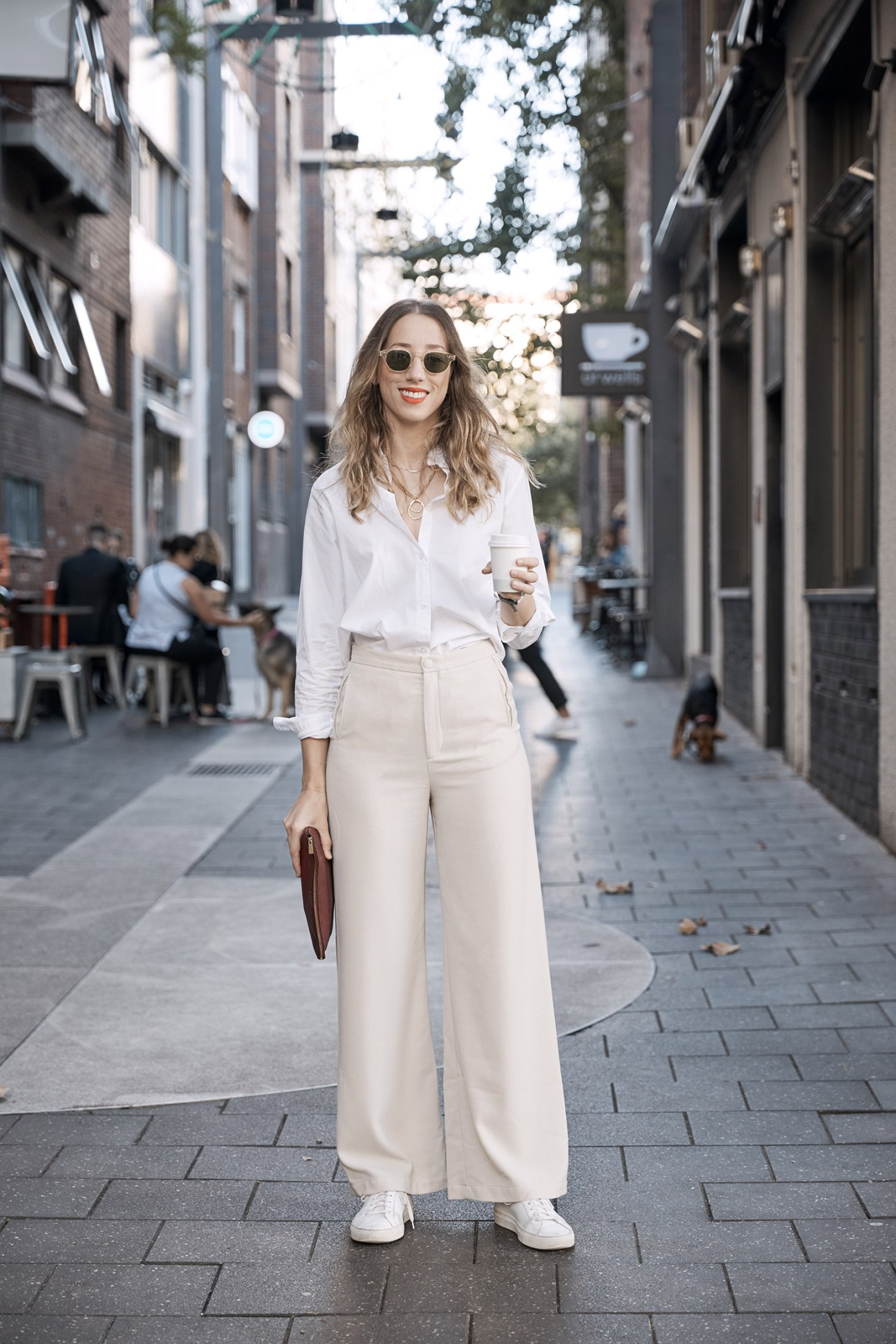 Woman in white shirt and cream trousers standing on urban street with coffee cup