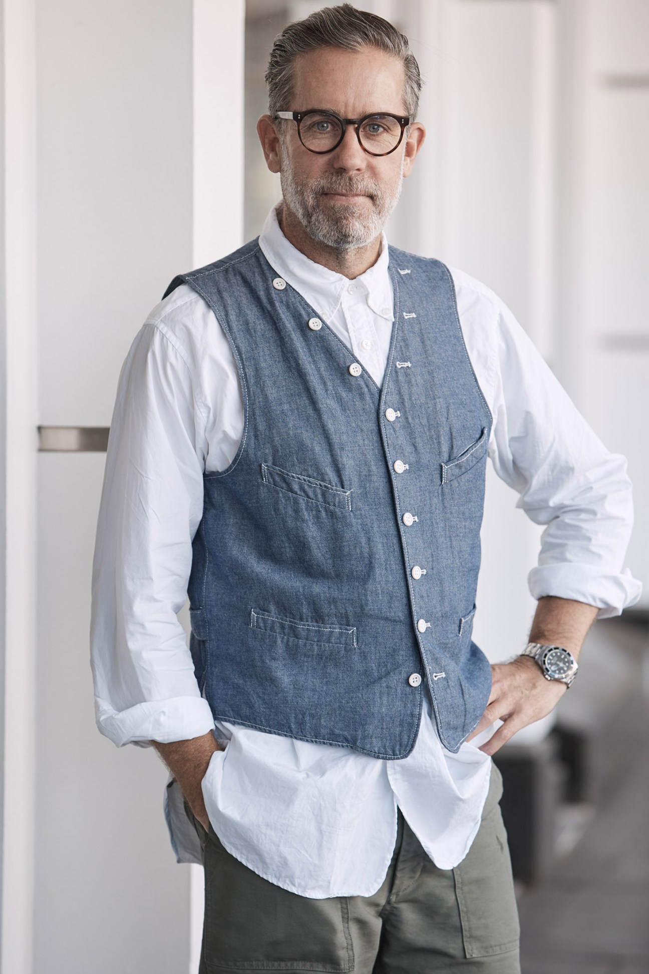 Man wearing glasses and denim vest over white shirt smiles at camera in indoor setting.