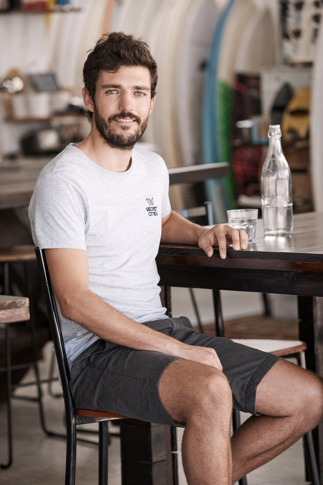 Man wearing light grey t-shirt with Secret Cities logo sitting at wooden table with water bottle and glass.