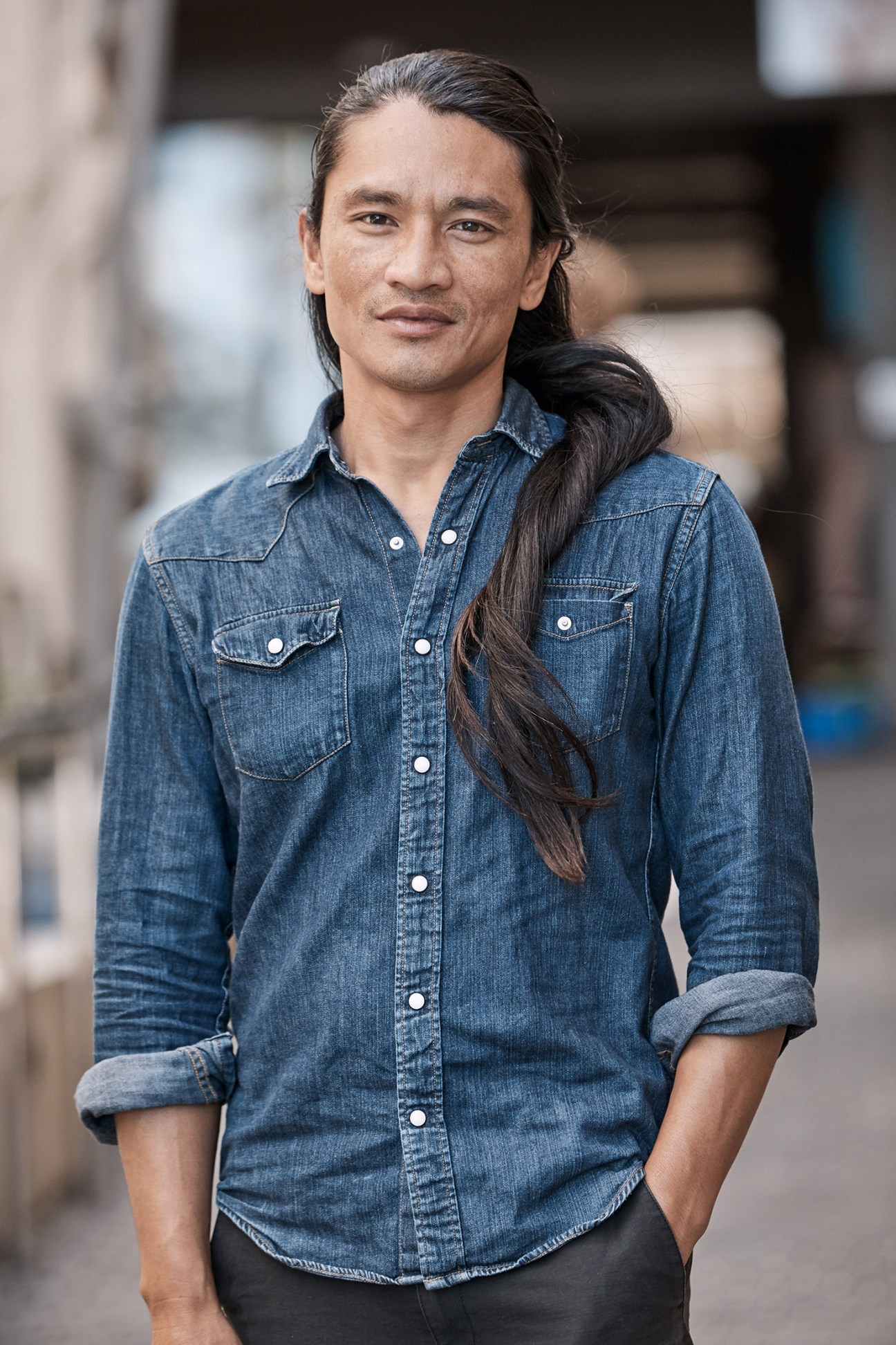 Man in denim shirt with long dark hair pulled back, standing outdoors in casual pose.