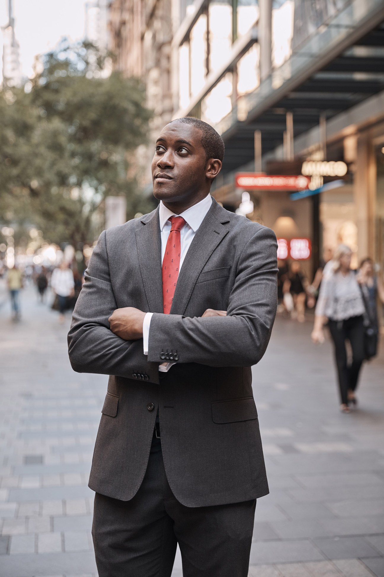 Man in grey suit and red tie standing on urban street with blurred pedestrians and buildings behind him.
