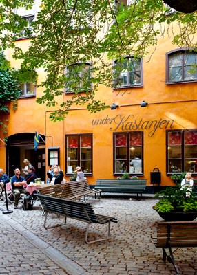 Orange building facade with ivy vines, outdoor seating area with benches and people gathered in a European courtyard