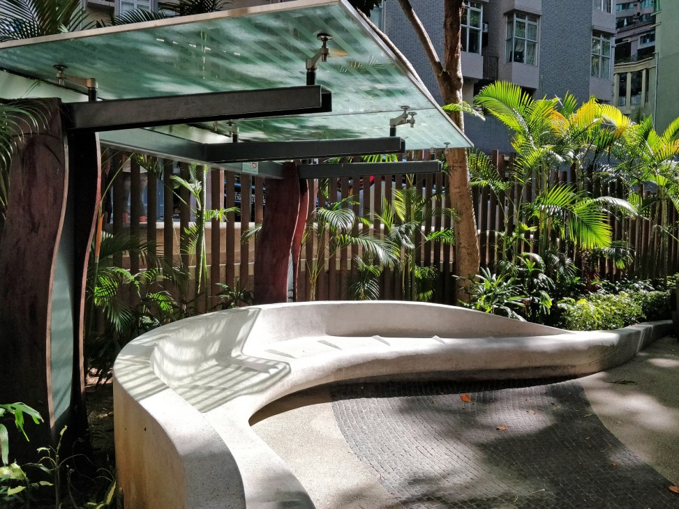 A curved white concrete bench with shade structure in a Hong Kong playground surrounded by tropical plants.