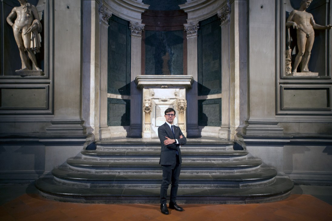 Man in dark suit standing in classical museum gallery with marble statues and ornate columns.