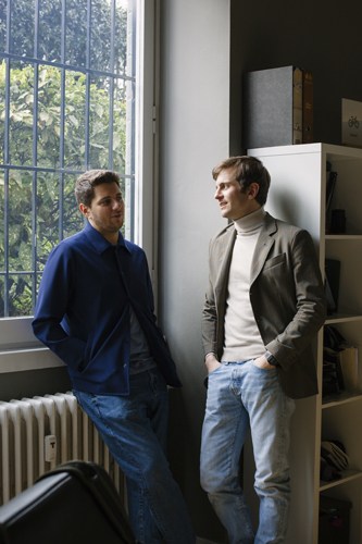 Two men in casual clothing stand in a bright studio space with large windows and shelving.