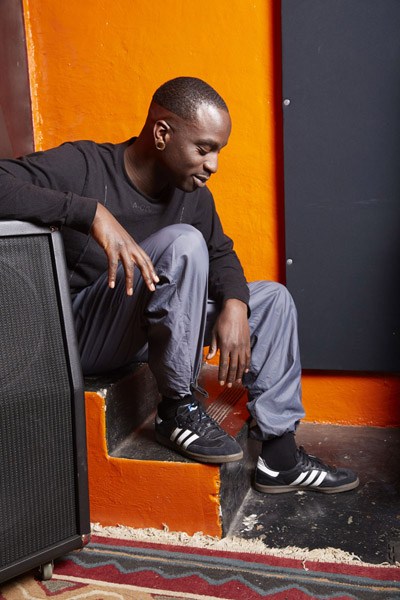 Man sitting on chair against orange and black wall wearing black shirt and gray pants