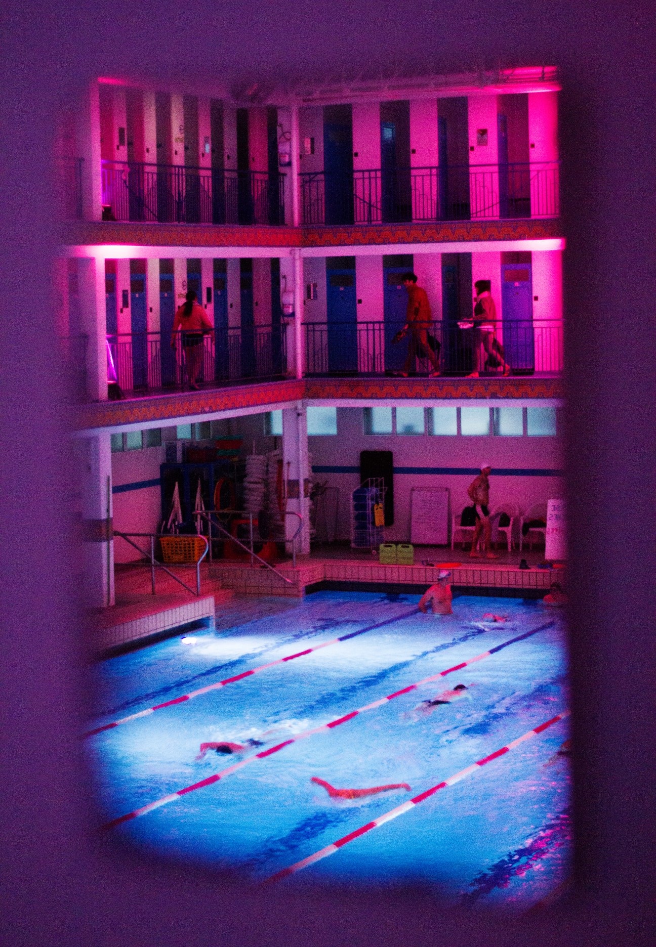 La Piscine de Pontoise illuminated at night with neon pink and blue lighting, showing swimmers in lanes and multi-story cabin galleries.