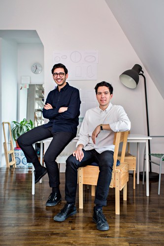 Two men pose in a bright studio space with wooden chairs, a potted plant, and a black floor lamp.