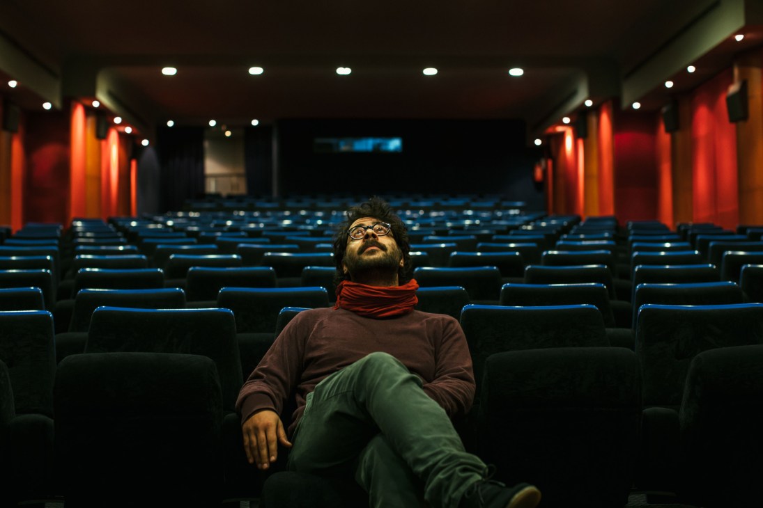 Man sitting alone in an empty movie theater with blue seats and red-orange wall panels.