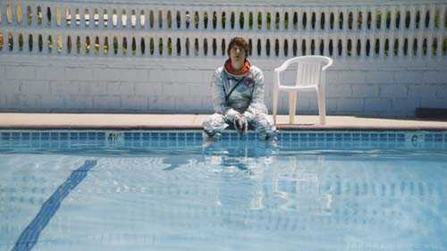Person sitting poolside in casual attire by water with white fence in background.