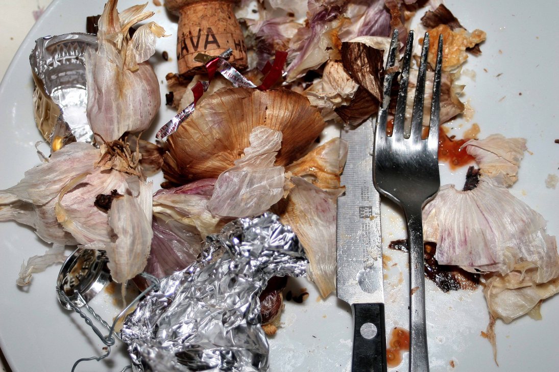 Overhead view of garlic cloves, foil wrappers, and a fork on a white plate with food remnants.