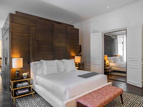 Hotel bedroom with dark wood headboard, white bedding, geometric patterned rug and orange bench