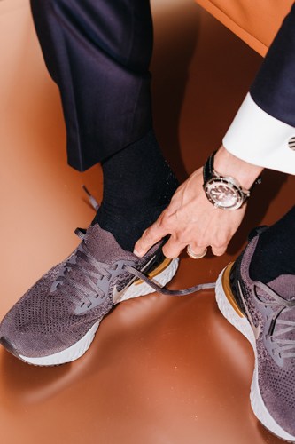 Manfred Fitzgerald wearing running shoes and a watch against an orange background.