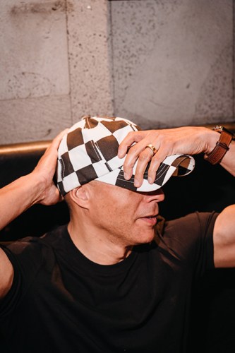 Man wearing brown and white checkered cap adjusting it with his hand while wearing a black shirt and ring.