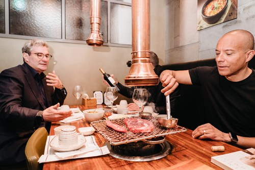 Two men dining at a Korean barbecue table with a central grill and copper hood.
