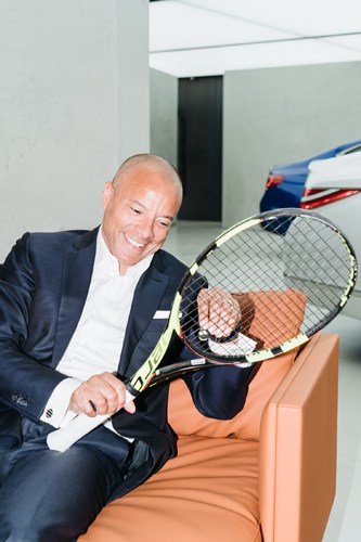 Manfred Fitzgerald holds a tennis racket while seated on an orange chair.