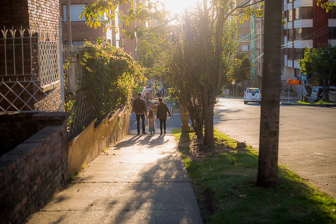 Pedestrians walking down a tree-lined street in Chapinero Alto, Bogotá, with brick buildings and dappled sunlight.
