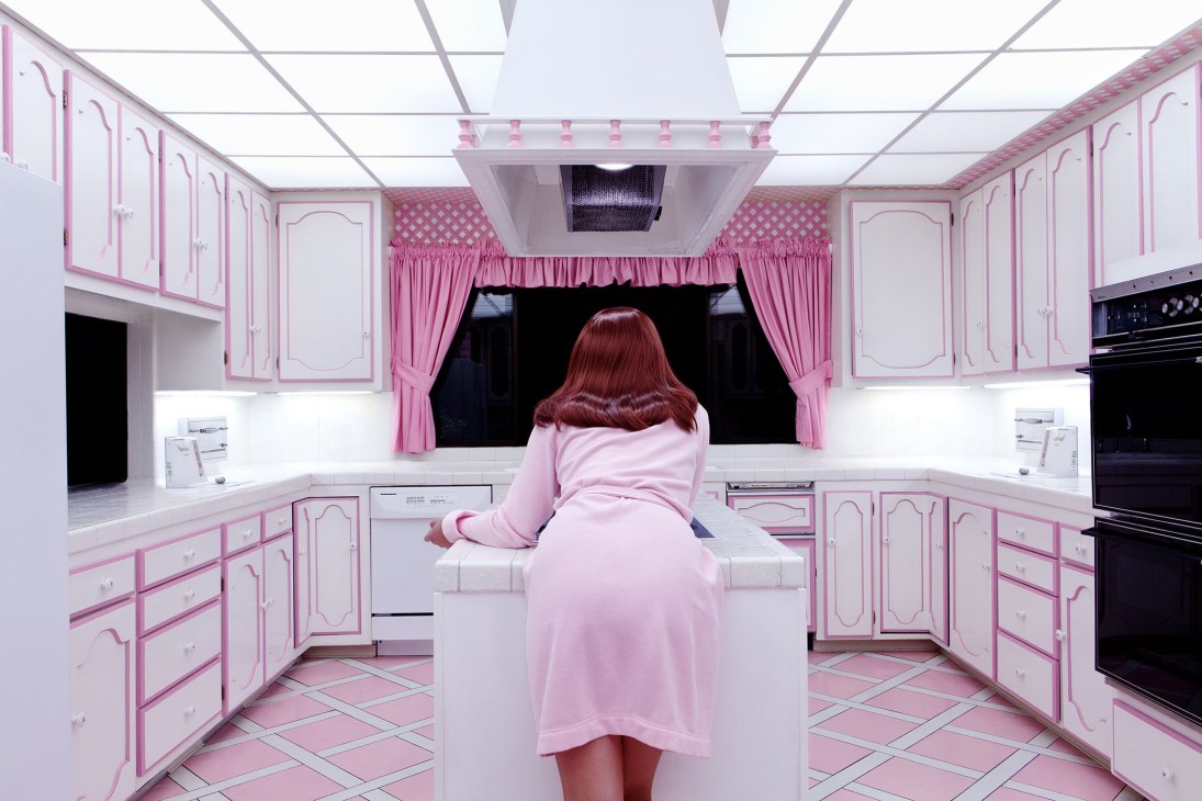 Woman with reddish hair viewed from behind in a pink and white retro kitchen interior.
