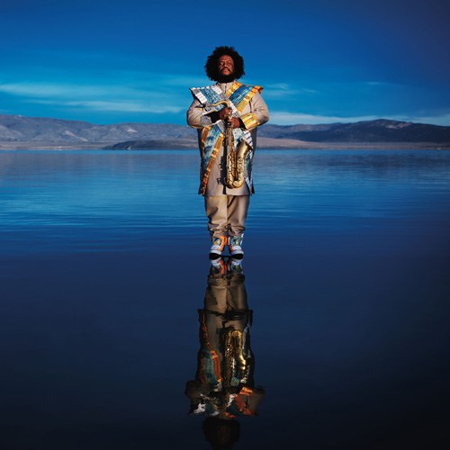 Kamasi Washington holding a saxophone while standing in water against a blue sky backdrop.